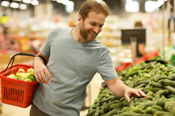 Happy customer standing and choosing green fresh cucumbers in section with vegetables in supermarket. Cheerful man smiling and holding red basket with products in hands.