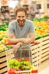 Front view of handsome bearded man posing with trolley in supermarket. Customer standing in section with citruces. Buyer leaning on trolley, looking at camera and smiling.
