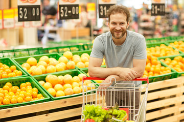 Handsome buyer standing in supermarket and leaning on trolley with products. Bearded man standing...