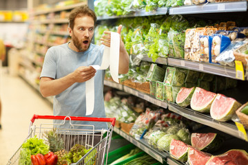 Bearded male customer standing near shelves with vegetables. Shocked man with wide opened eyes looking at long grocery receipt. Trolley full of products placed near buyer.