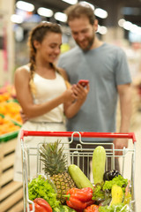 Close up of trolley full of fresh colorful vegetables. Happy couple standing on background. Woman and man smiling and looking at phone. Concept of trade and shopping in vegetable section.