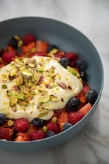 Beeren Obstsalat mit Erdbeeren, Himbeeren, Blaubeeren, Pistazien, Zitronen Zesten, Leinöl Kurkuma Quark Creme in blauer Schale als Frühstück auf Marmor Hintergrund