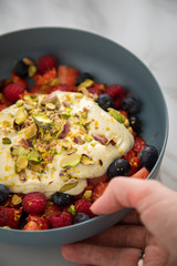 Frau isst Beeren Obstsalat mit Erdbeeren, Himbeeren, Blaubeeren, Pistazien, Zitronen Zesten, Leinöl Kurkuma Quark Creme in blauer Schale als Frühstück auf Marmor Hintergrund