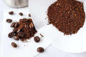 Dark roasted coffee beans and ground coffee in white paper filter on white cotton fabric table top in natural light