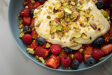 Beeren Obstsalat mit Erdbeeren, Himbeeren, Blaubeeren, Pistazien, Zitronen Zesten, Leinöl Kurkuma Quark Creme in blauer Schale als Frühstück auf Marmor Hintergrund