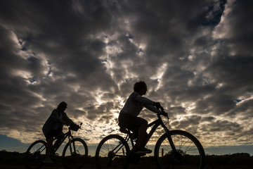 Obraz premium Couple of woman riding mountain bike in silhouette with great beautiful dramatic clouds sky in background - epic image for outdoor sport leisure activity