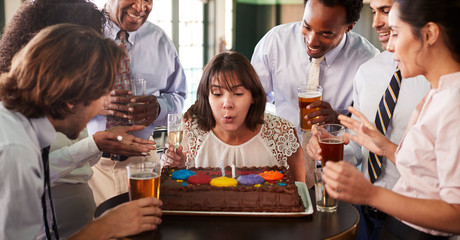 Businesswoman Blowing Out Candles On Birthday Cake At Celebration In Bar With Colleagues