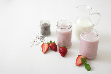 Strawberry smoothie, milkshake. 2 jars of strawberry milk smoothie and 1 jar of chia seeds on white table, embellished with real strawberries and fresh mint. Glass pitcher with milk in background