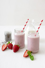 Strawberry smoothie, milkshake. 2 jars of strawberry milk smoothie and 1 jar of chia seeds on white table, embellished with real strawberries and fresh mint. Glass pitcher with milk in background