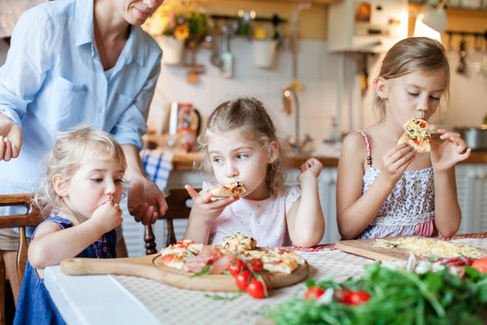 Children are eating and tasting italian homemade pizza cooking themselves together. Cute kids are enjoying delicious food in cozy home kitchen. Three girls at family dinner table. Lifestyle moment.