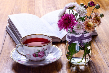 Tea break time. Tea cup, wildflowers in vase and open book on wooden background.