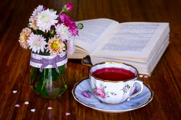 Tea break time. Tea cup, wildflowers in vase and open book on wooden background.