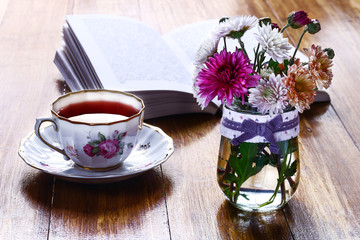 Tea break time. Tea cup, wildflowers in vase and open book on wooden background.
