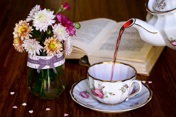 Tea break time. Tea cup, tea pot, wildflowers in vase and open book on wooden background. Teapot...