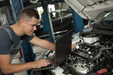 View from side of concentrated man in process of doing diagnostacs of vehicle at car service. Proffesional mechanic leaning on open hood of car, and working with computer. Concept of car checking.