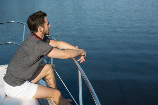 Side view of bearded man sitting on boat bow, thinking, dreaming and looking away at sea. Man wearing white shorts and grey t shirt. Concept of yach, sail boating and voyage. - Powered by Adobe
