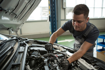 Fototapeta na wymiar Male mechanic leaning over open hood, keeping wrench and fixing problem in car. Qualified man in grey uniform and protective gloves repairing automobiles in car service. Concept of maintenance.