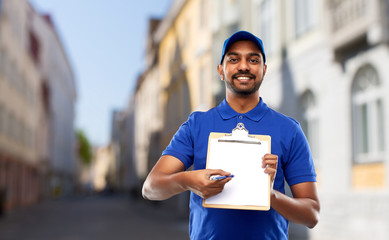 mail service and shipment concept - happy indian delivery man with clipboard in blue uniform over old tallinn city street background