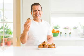 Middle age man eating chocolate chip muffins at home with a happy face standing and smiling with a...