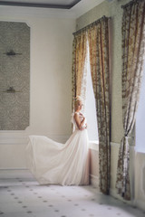 Cute blonde girl in the image of the bride stands near the window in a flying bright dress. wedding photo. gentle calm image. Small Noise