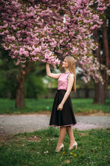 Attractive girl in dress stand in park by the blossom tree. Blond hair female