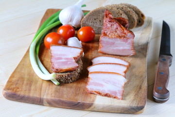 A piece of bacon on a chopping board with vegetables and bread.