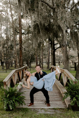 Fototapeta premium Beautiful Bridesmaid Woman in Blue Dress and Bouquet with Her Date at a Formal Wedding Party Celebration Event Outside in the Woods Taking Couple Portraits on Small Wooden Bridge