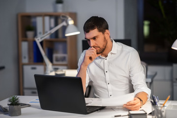 business, deadline and people concept - businessman with laptop computer and papers working at night office