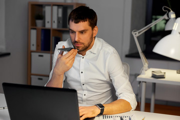 business, deadline and technology concept - businessman with laptop computer using voice command recorder on smartphone at night office