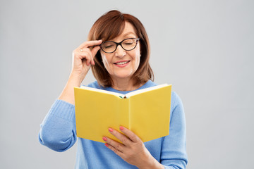 vision, wisdom and old people concept - portrait of smiling senior woman in glasses reading book over grey background