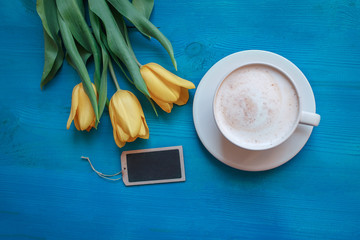 Coffee mug with yellow tulip flowers and notes on blue rustic table from above, breakfast on Mothers day or Womens day