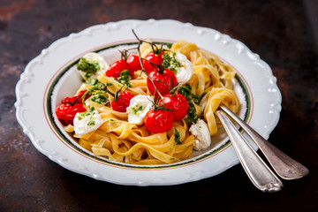Fettuccine pasta with cherry tomatoes, mozzarella and pesto sauce. Hot dish with baked vegetables on a dark background.