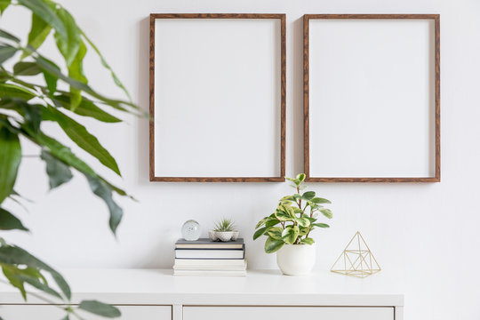 Stylish home interior with two brown wooden mock up photo frames on the white shelf with books, beautiful plants, gold pyramid and home accessories. Minimalistic concept of white room decor. 