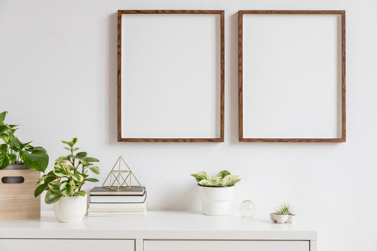 Elegant home interior with two brown wooden mock up photo frames above the white shelf with books, plants, gold pyramid, wooden box and home accessories. Stylish concept of white room decor. 