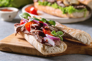 Beef baguette sandwich with tomatoes, basil, red onion served on ceramic plate with ingredients above over grey blue table.