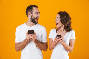 Image of excited couple man and woman smiling while both using mobile phones, isolated over yellow...