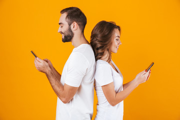 Image of modern couple man and woman smiling while both using mobile phones, isolated over yellow background