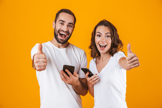 Image of content couple man and woman smiling while both using mobile phones, isolated over yellow background