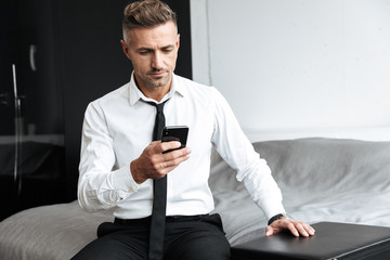 Business man in bedroom indoors at home sitting on bed using mobile phone.