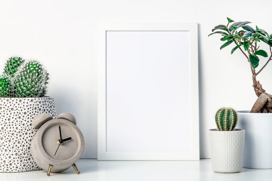 White shelf at home. Cactus and bonsai decoration in concrete and ceramic pots. Concrete clock. White empty frame mockup. Space for text or graphics