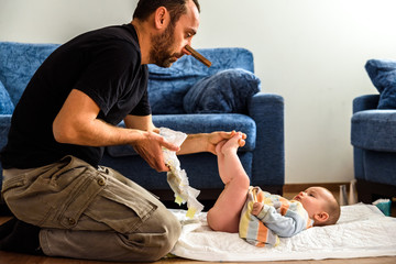 Dad with a pincer on his nose so he does not smell the smell of his daughter's diaper while he cleans and changes her, fatherhood. - obrazy, fototapety, plakaty
