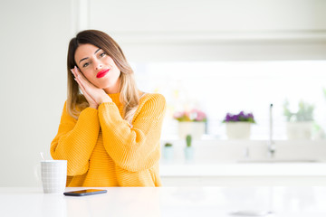 Young beautiful woman drinking a cup of coffee at home sleeping tired dreaming and posing with hands together while smiling with closed eyes.