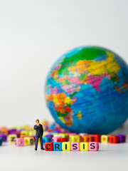 miniature figure businessman in dark blue suit standing backside of colorful of CRISIS alphabet and globe in the background and thinking of the crisis will come.