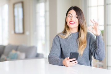 Young beautiful woman using smartphone at home doing ok sign with fingers, excellent symbol