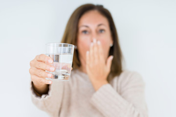 Middle age woman drinking glass of water isolated background cover mouth with hand shocked with shame for mistake, expression of fear, scared in silence, secret concept