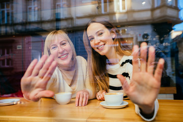 View through the window of blonde adult girl with her mature mother drinking coffee, they are sittting at cafe, waving you. Bonding, different generationsconnect, family concept.