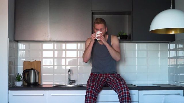 Young Ginger Man drinking coffee and sitting on a counter top in kitchen
