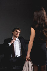 brunette woman standing opposite handsome man sitting on couch and taking off tie on dark background