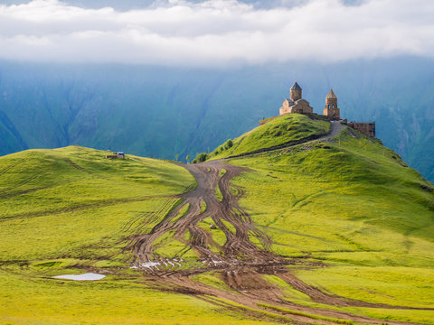 Colorful horizontal photo of Gergeti Tsminda Sameba church in Kazbegi (Stepancminda) village, Georgia, Caucasus, on a spring morning. Juicy green grass covers the hills, white clouds above the church.