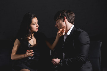 attractive brunette woman in black dress and handsome man in suit sitting on couch with glass on black background
